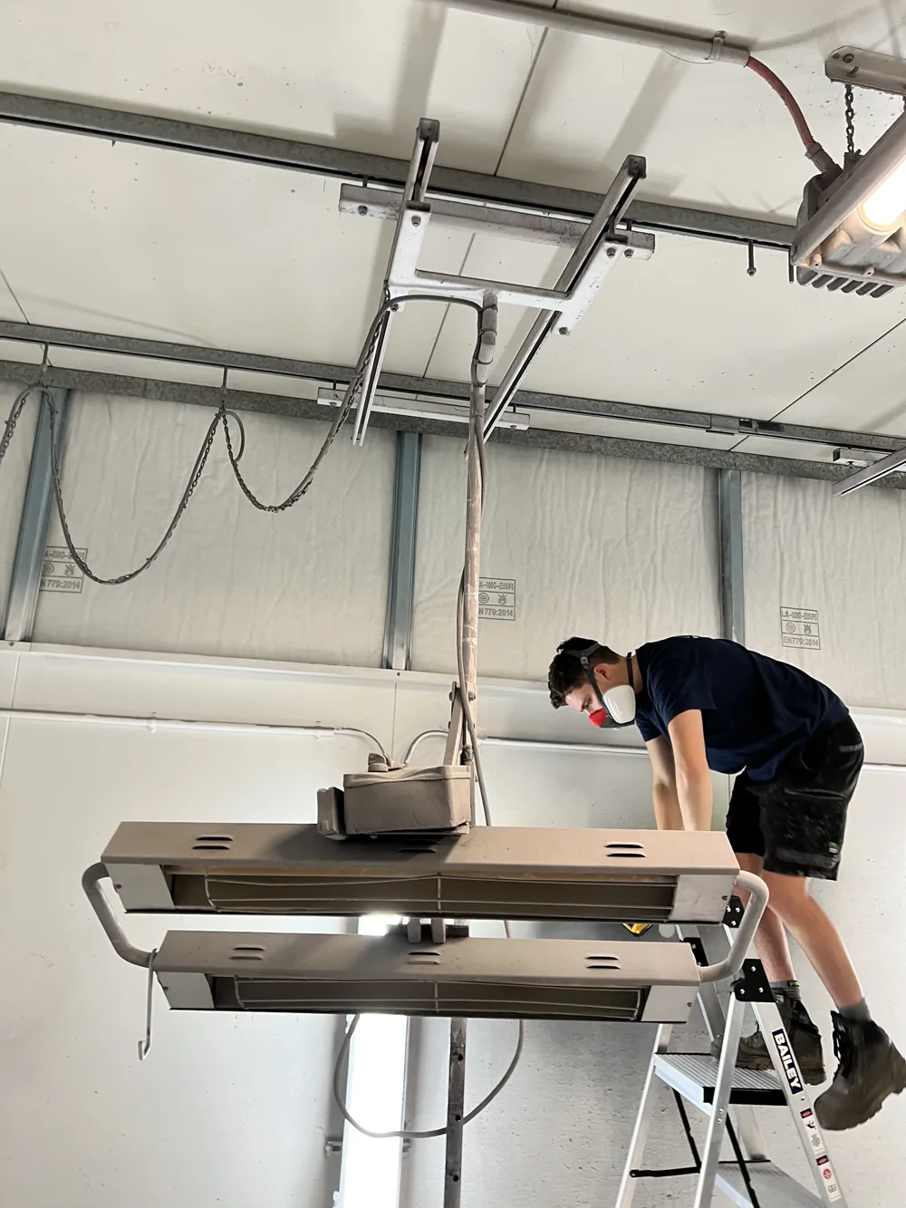 Worker wearing a safety mask on a ladder, repairing or maintaining overhead industrial equipment.