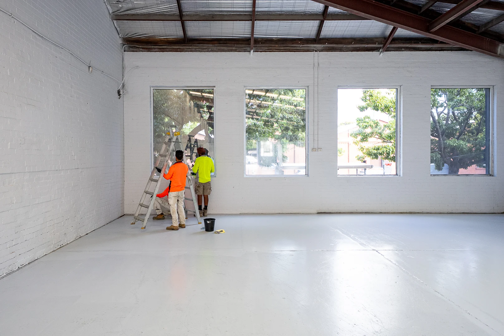 Workers on a ladder cleaning or installing glass on large windows inside a bright industrial space.